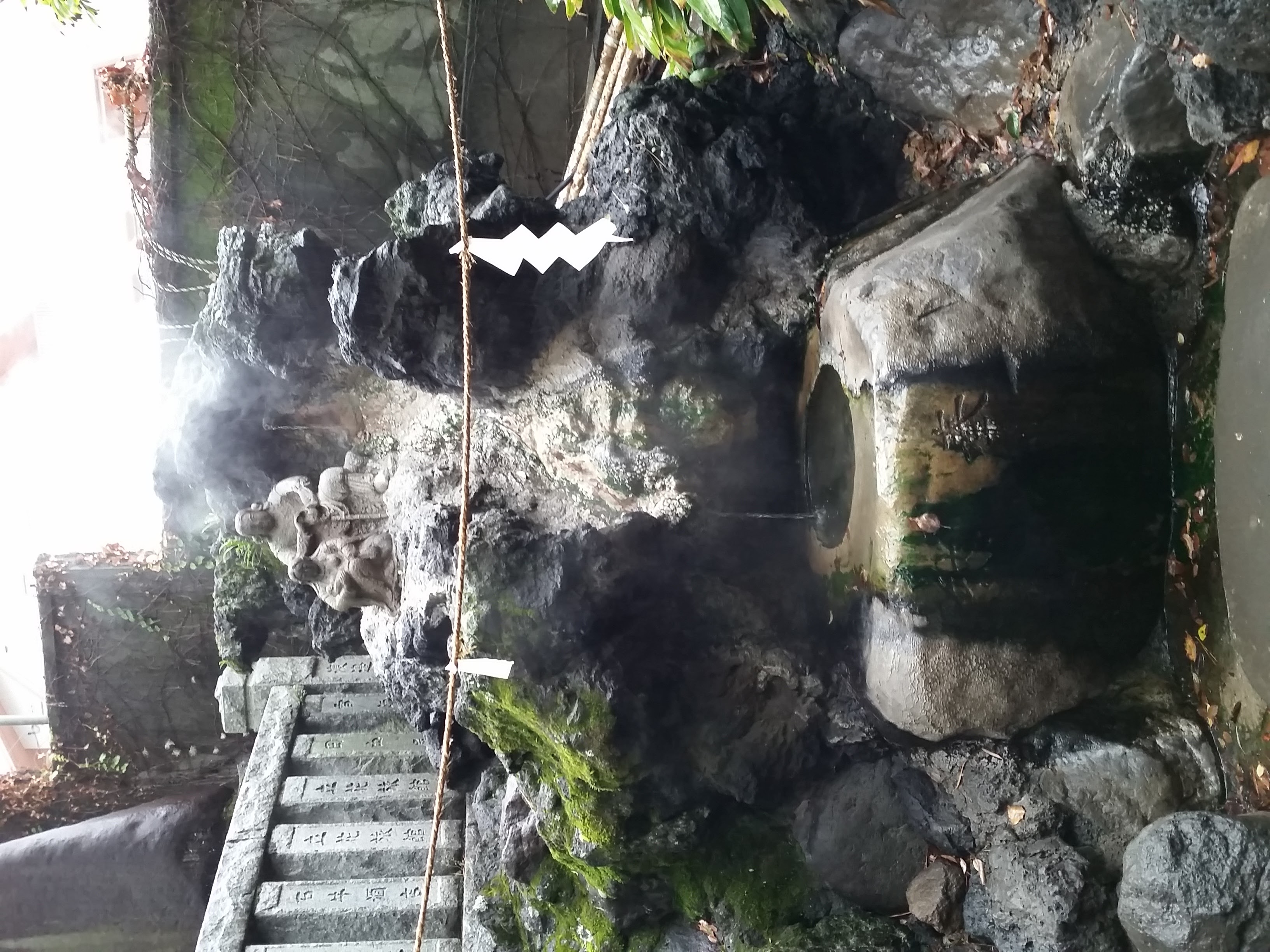 Natural onsen spring in Atami with steam rising from moss-covered rocks, shimenawa rope and stone steps with inscriptions