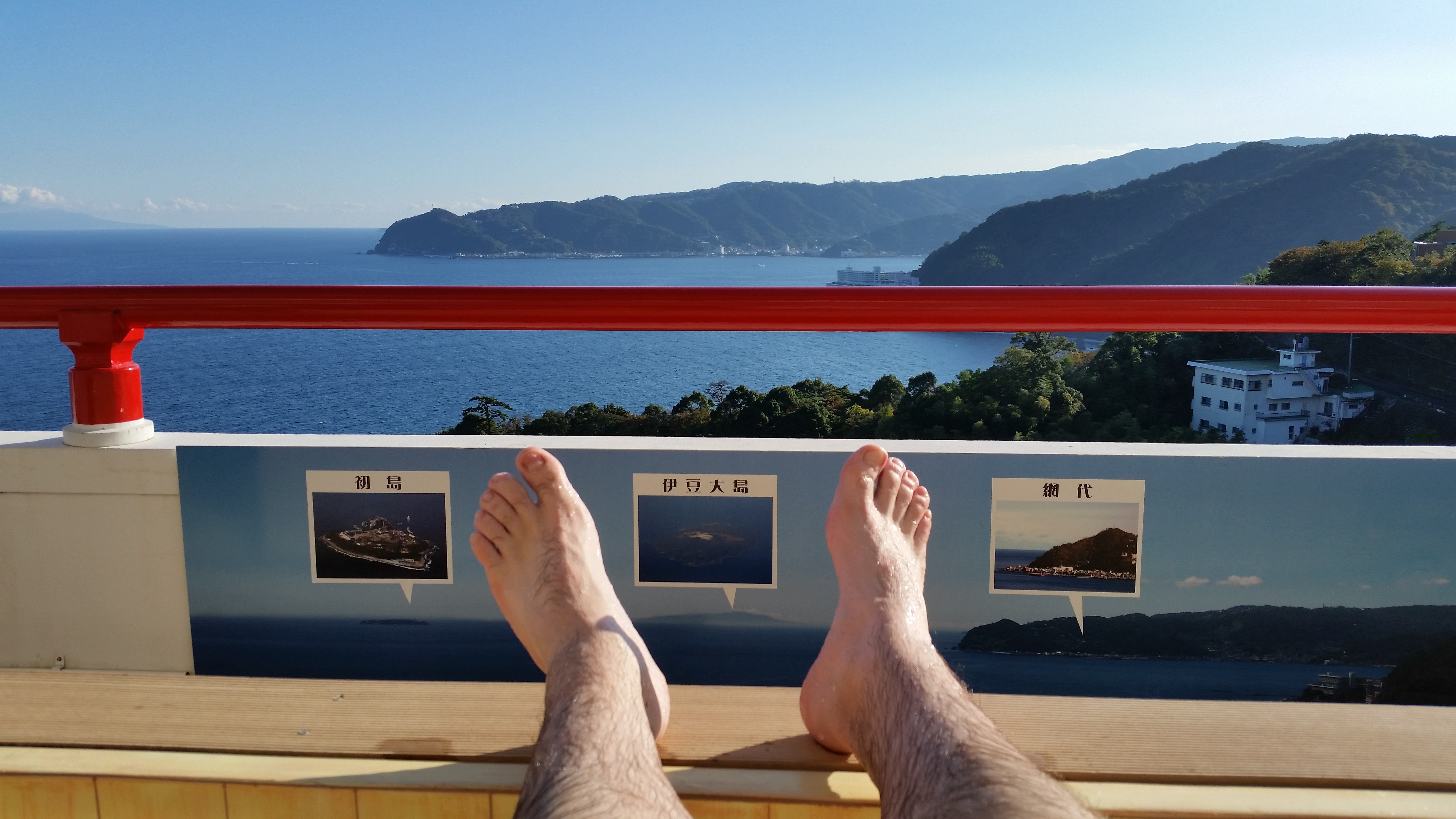 Bare feet resting on the edge of a ryokan rotenburo with views of Sagami Bay and the outlines of Hatsushima and Izu Oshima islands on the horizon