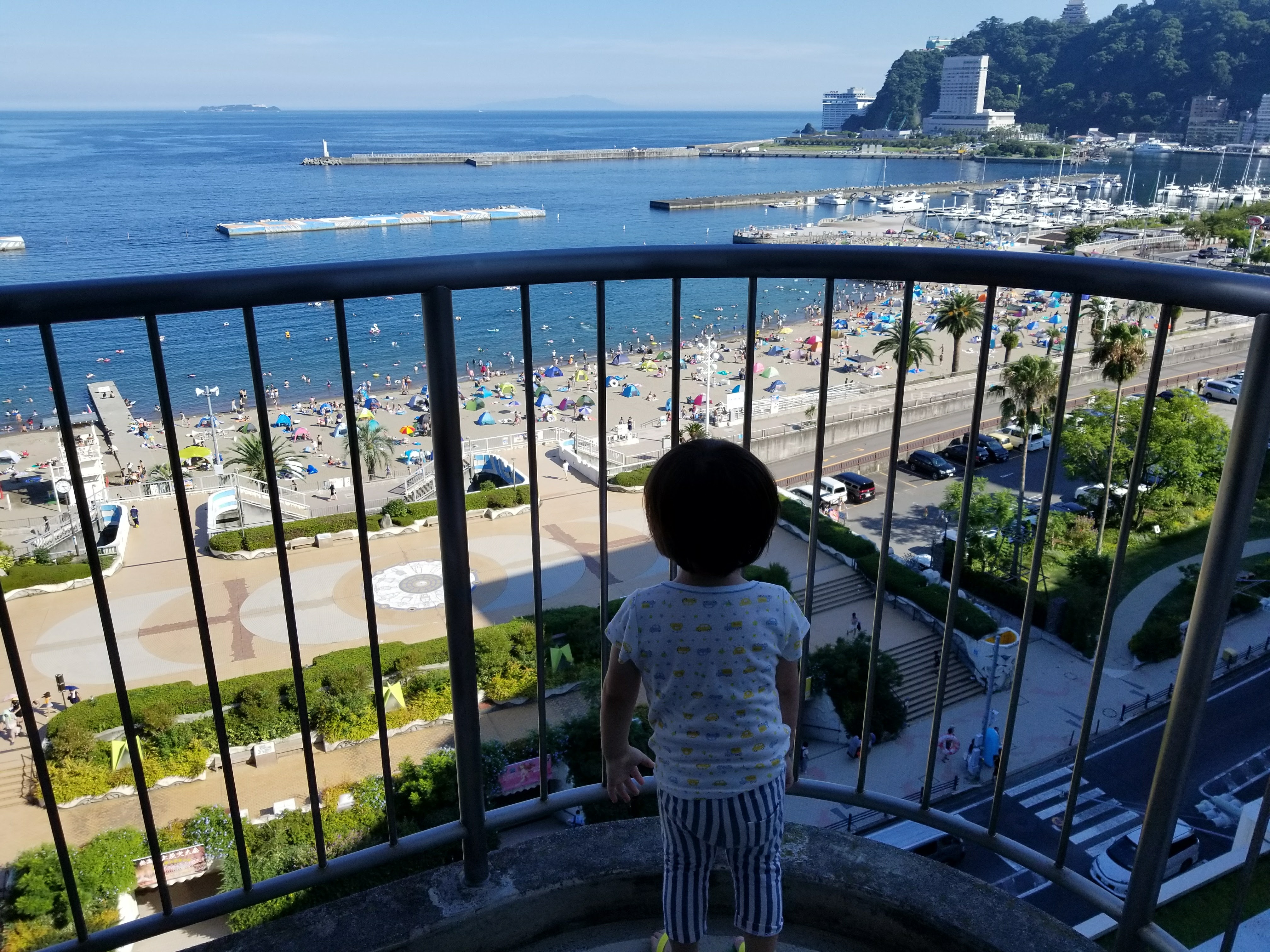 Child standing at a ryokan balcony looking out at Atami beach packed with swimmers and colourful umbrellas, with the marina visible in the background