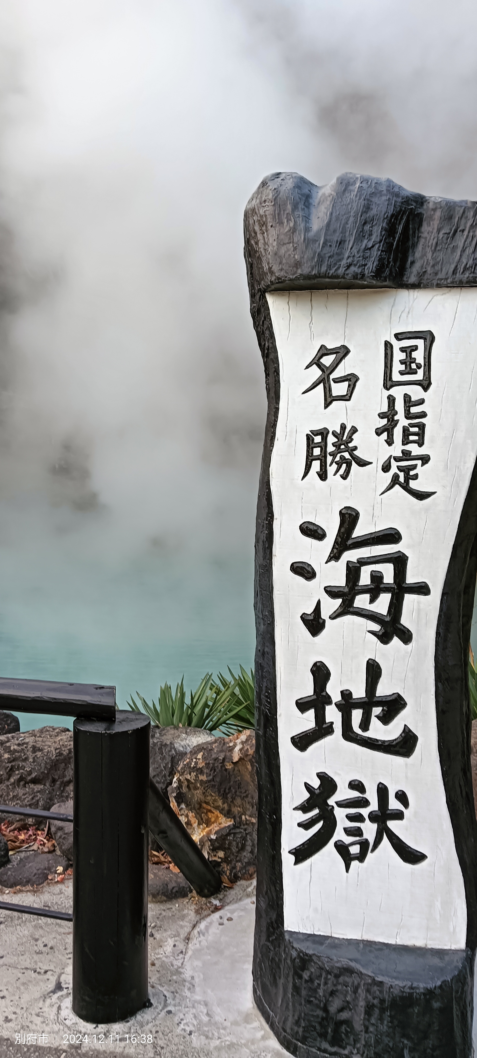 Wooden sign for Umi Jigoku with the kanji "海地獄" carved in black, with the intensely turquoise thermal water and steam visible behind it