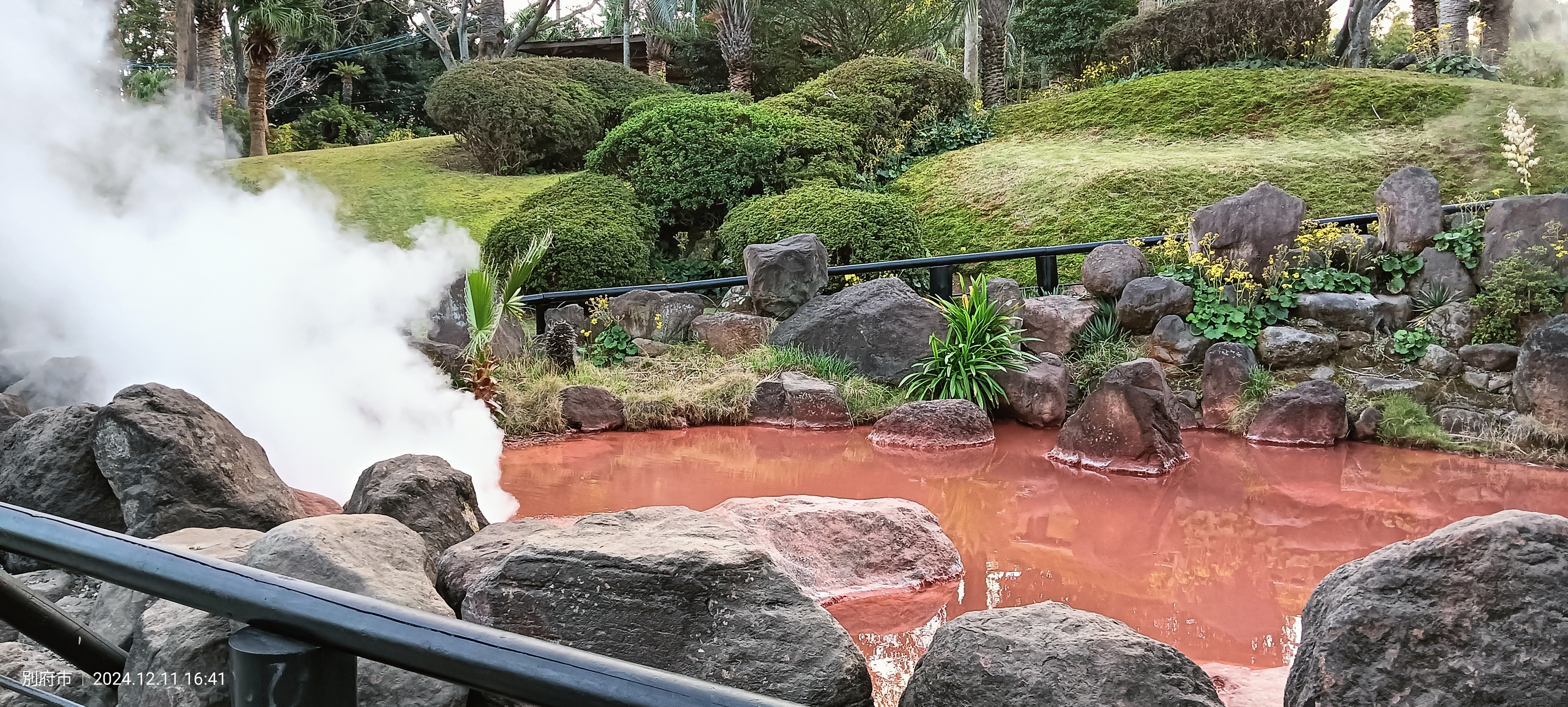 View of Chi-no-ike Jigoku with the intensely red water between the rocks and the green garden in the background