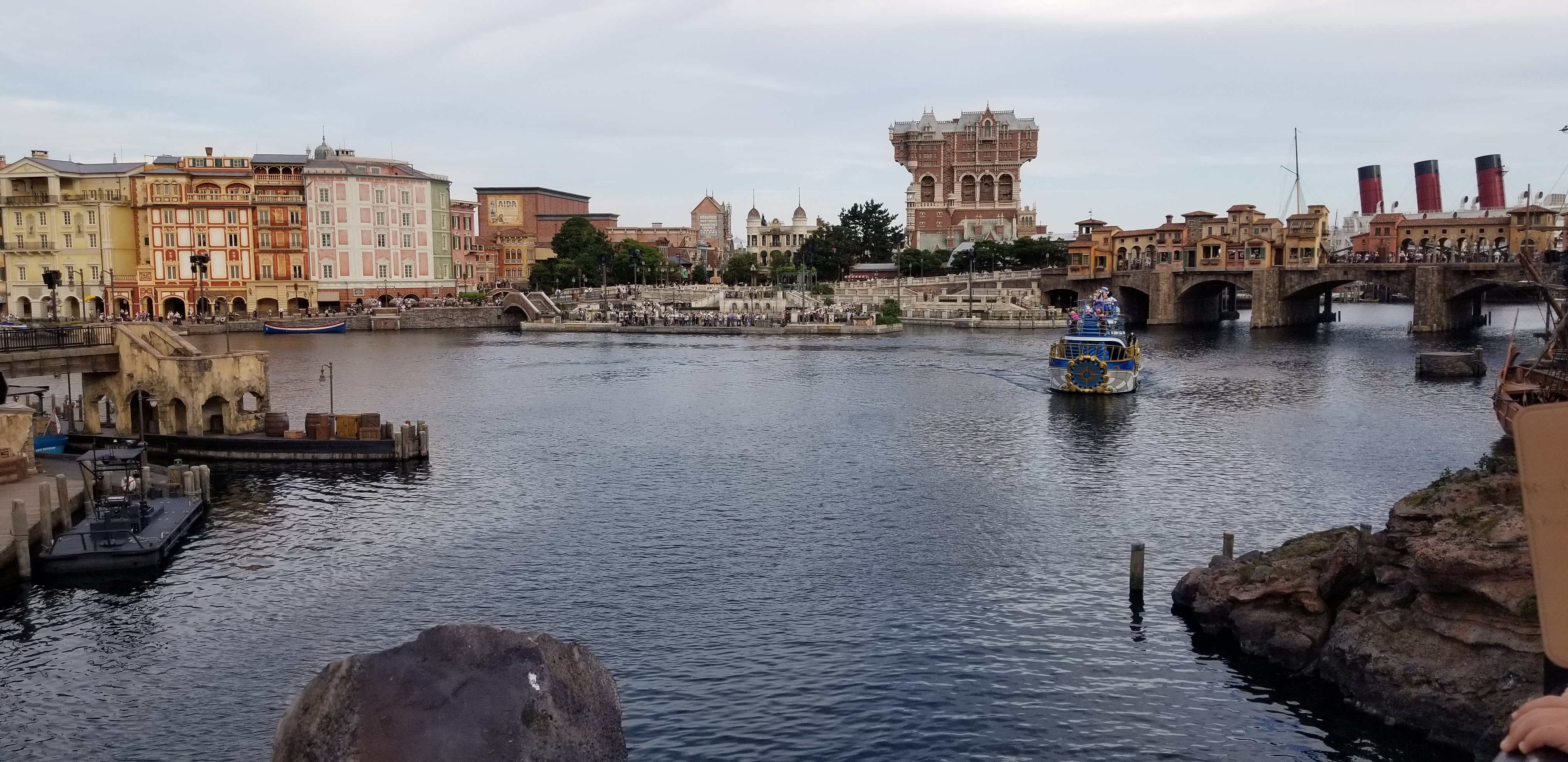 Panoramic view of DisneySea's Mediterranean Harbor with Italian-style buildings, the Tower of Terror in the background and the parade ship on the water