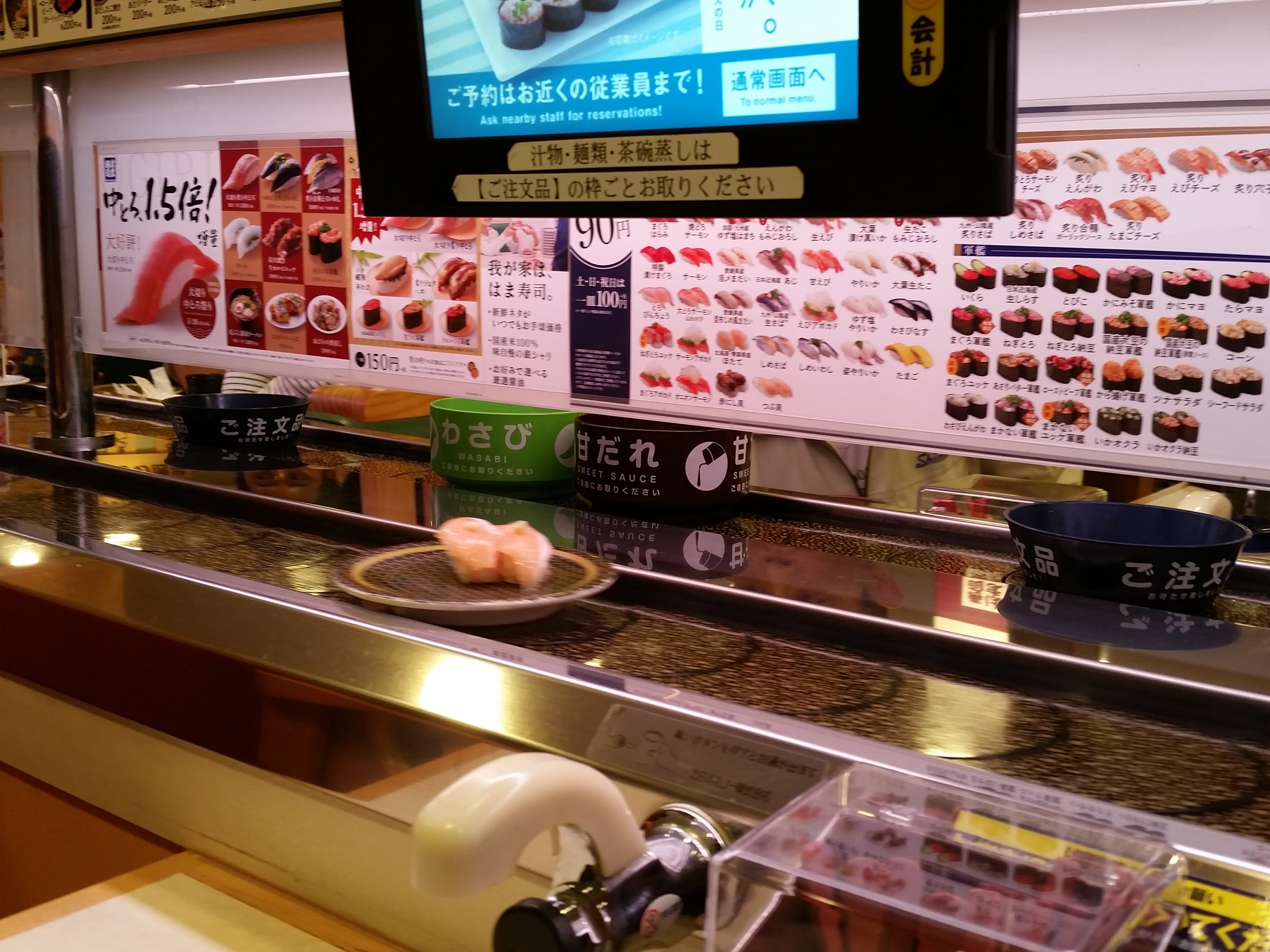 La cinta transportadora de un Hama Sushi con platos en movimiento, la pantalla de pedidos táctil y los dispensadores de wasabi y salsa dulce